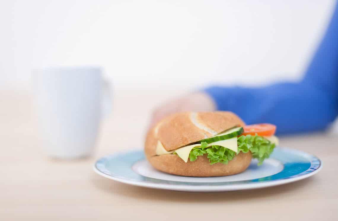 Ein belegtes Brötchen mit Käse und Tomaten, im Hintergrund eine frau mit blauem Pullover und eine weisse Tasse: passend zum Thema: Kommt Dein Brötchen aus China?