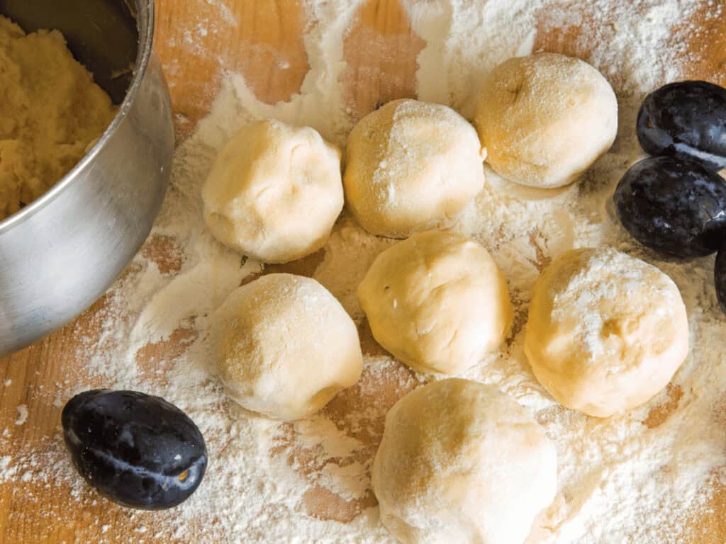 Ungekochte glutenfreie Zwetschgenknödel auf Mehl, daneben liegen Zwetschgen.