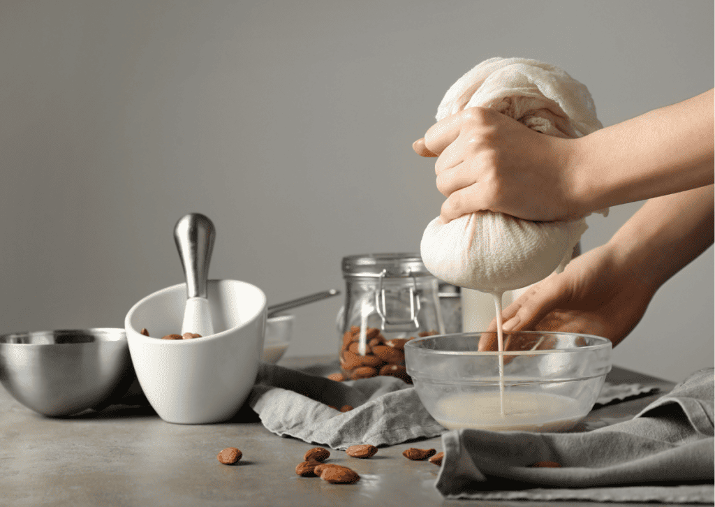 Auf dem Bild sind die Hände einer Frau, die histaminarme, glutenfreie Nussmilch machen. Sie presst die Nussmilch durch einen Nussmilchbeutel, im Hintergrund sind die Utensilien wie ein Glas mit Mandeln.