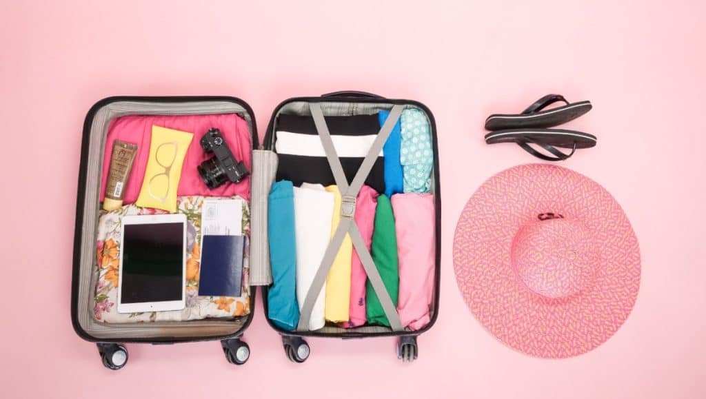Ein Koffer mit dem nötigsten Reiseutensilien wie kleidung, Brille Laptop, Sonnencreme und Kamera. Neben dem Koffer liegen Flip-Flops und ein rosa Sonnenhut. Stressfreier Reisen, bedeutet auch, kleineres Gepäck zu haben.